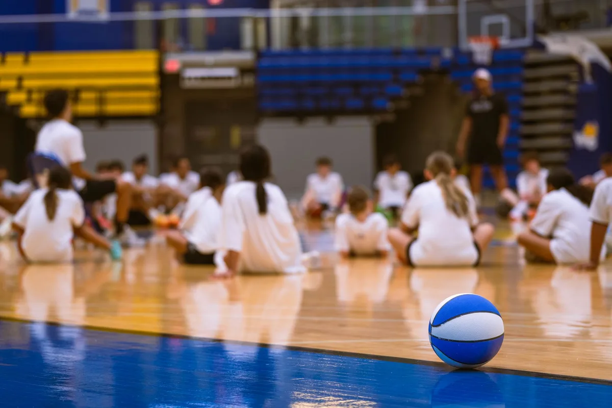 Coach in silhouette standing at the sideline of an empty high school gymnasium, facing the court with hands on hips
