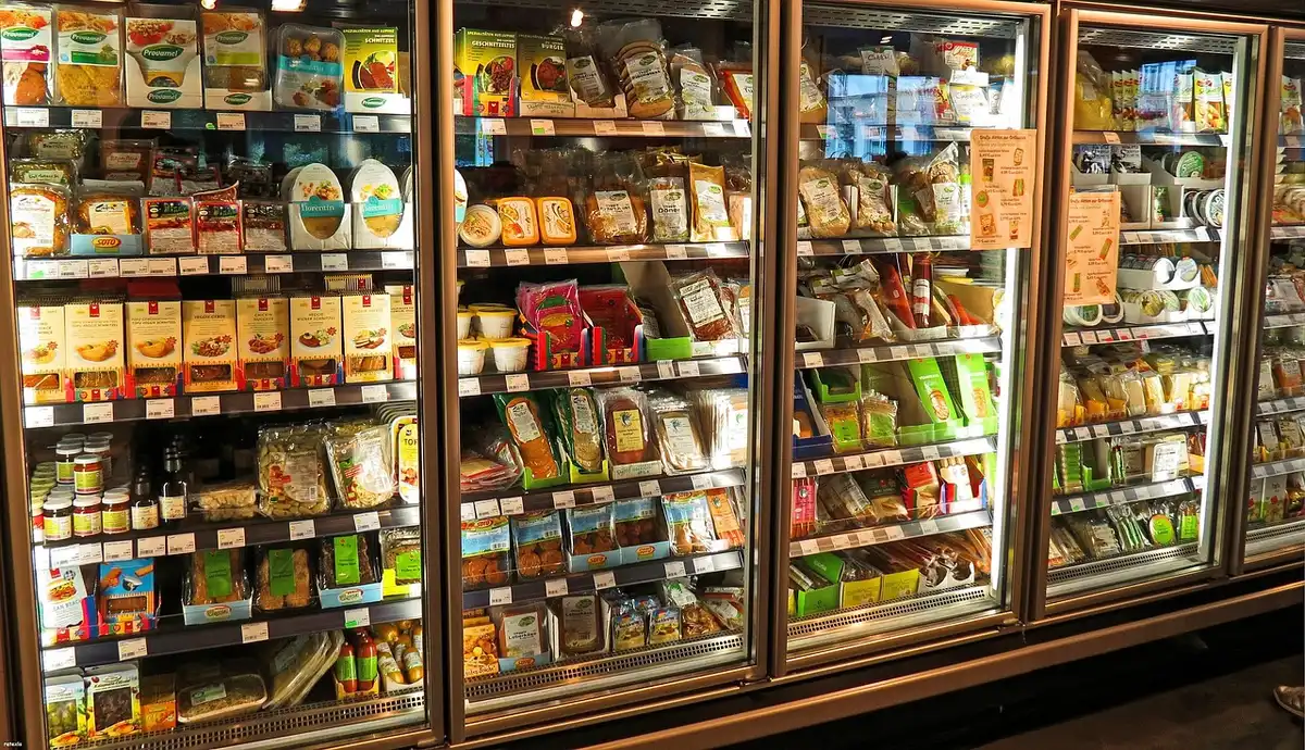 Shoppers reaching for frozen pizza boxes in a bright supermarket freezer aisle