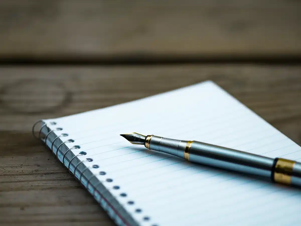 Bright desk with open notebook, pen and laptop in daylight, hands over notes